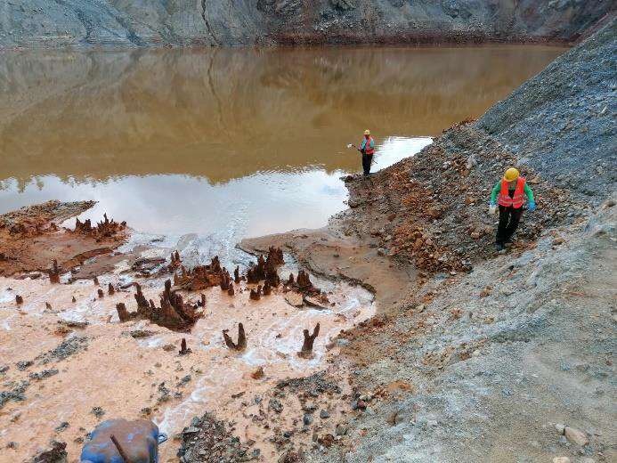 A pond of orange-brown water at the foot of a grey waste dump; two workers in high-visibility vests take a sample.